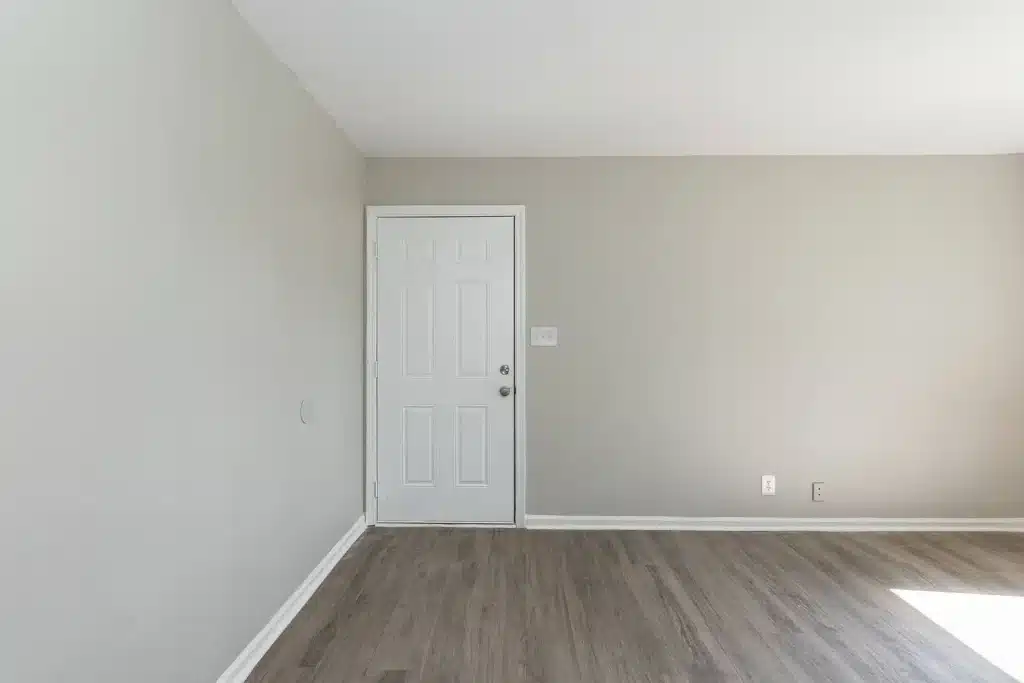 A white door with a silver handle is set in a light gray wall in a mostly empty room with wood-style flooring and white baseboards.