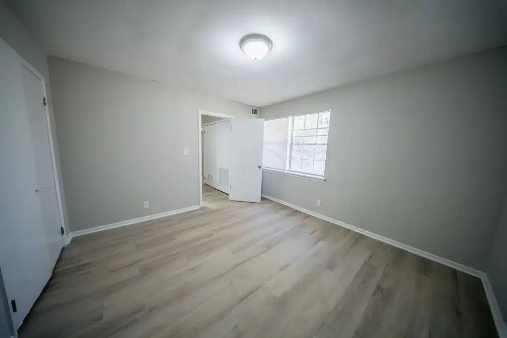 Empty room with light gray walls, wood-style flooring, a ceiling light fixture, a window with blinds, and two white doors, one open and one closed.