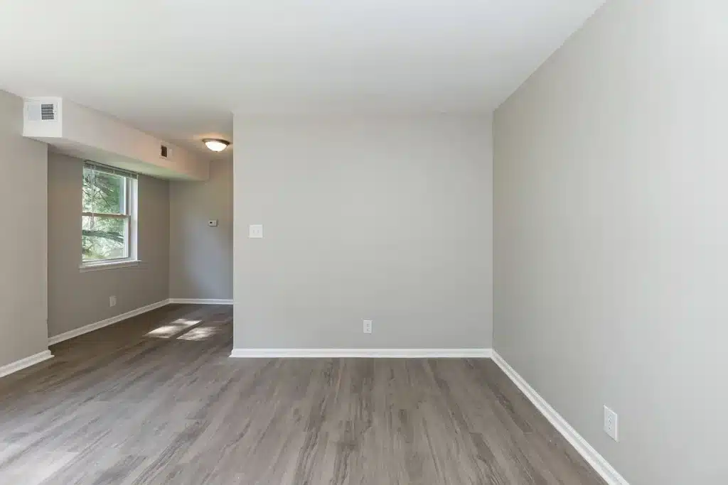 Empty room with light gray walls, wood-style flooring, a window on the left, and a small hallway leading to another area.