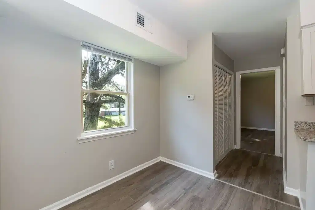 A small room with light gray walls, a window with blinds, wood-style flooring, and a hallway with white closet doors leading to another room.