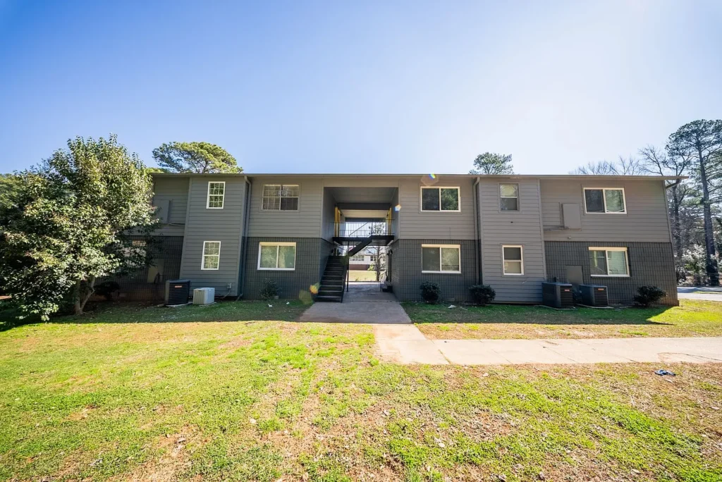 Two-story apartment building with gray siding, central staircase, and a grassy lawn in front under a clear blue sky.