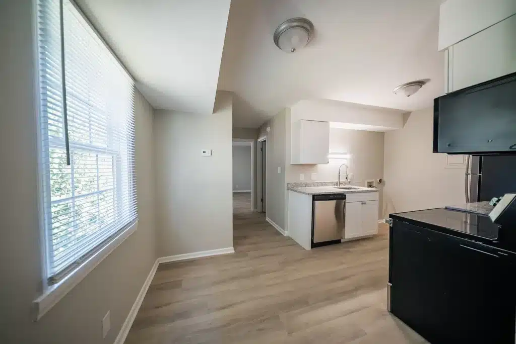 A small, modern kitchen with light wood floors, white cabinets, stainless steel appliances, and a window letting in natural light.