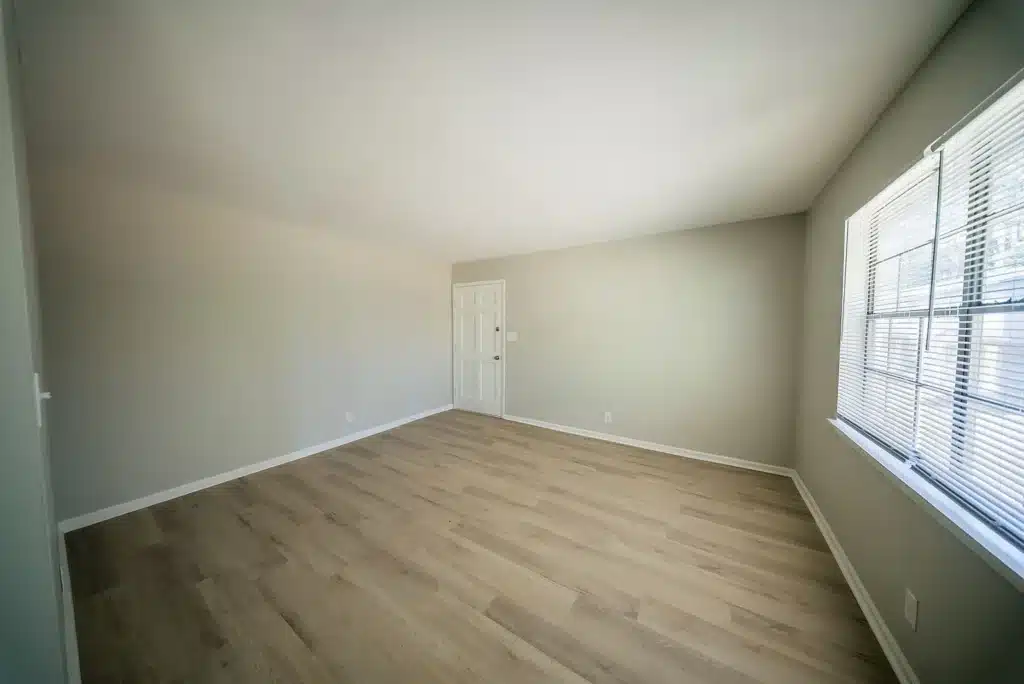 Empty room with light-colored walls, wood flooring, a closed white door, and a large window letting in natural light.