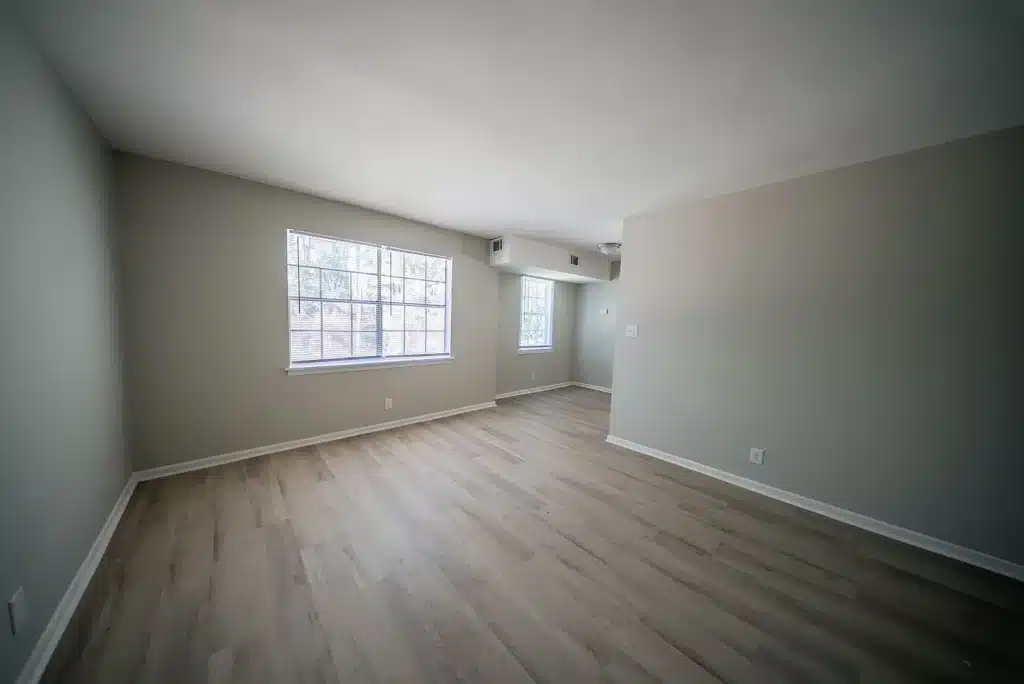 Empty living room with light wood flooring, gray walls, large window on the left, and a small dining area visible in the back right.