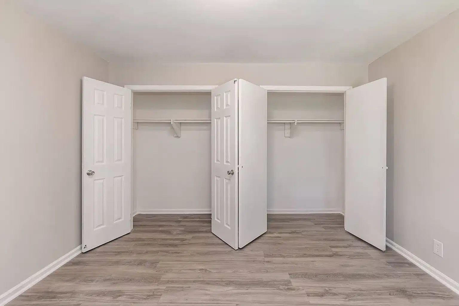 Empty double closet with open white doors, a single hanging rod and shelf in each section, light gray walls, and wood-style flooring.