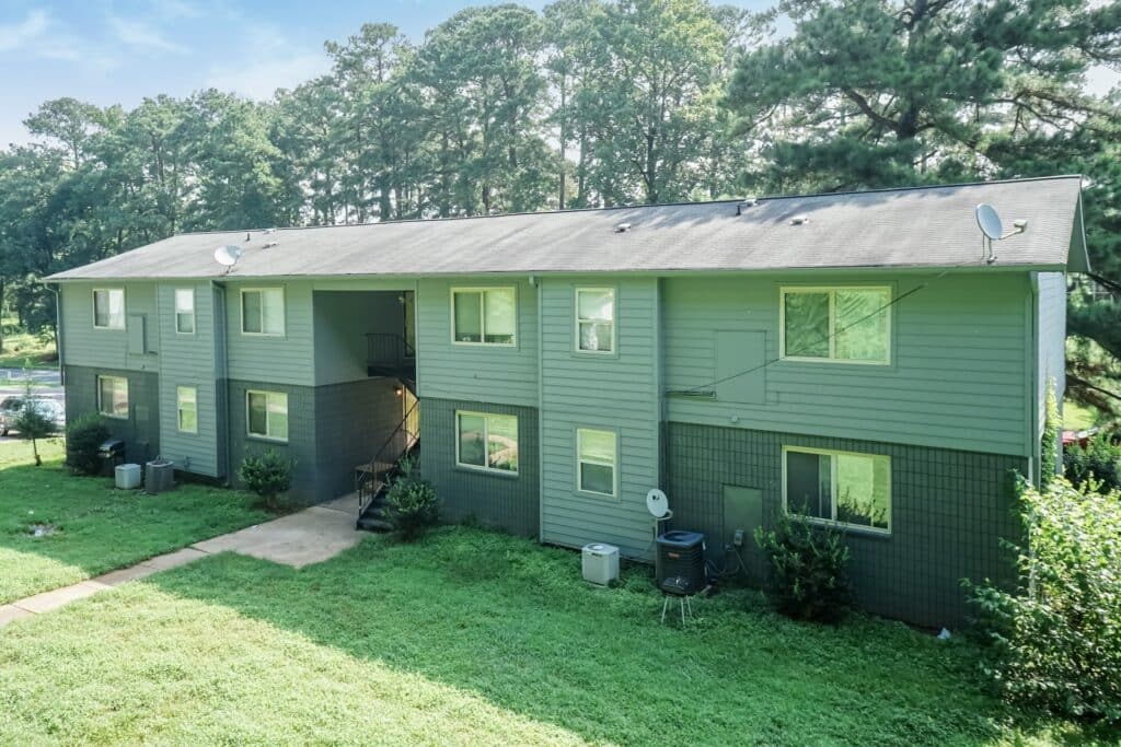 A two-story green apartment building with multiple windows and satellite dishes, surrounded by grass and trees, under a clear sky.