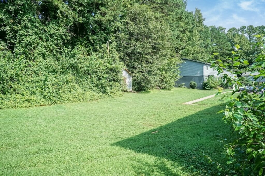 A green grassy yard bordered by dense trees and bushes, with a small shed in the back and part of a gray house and driveway visible on the right.