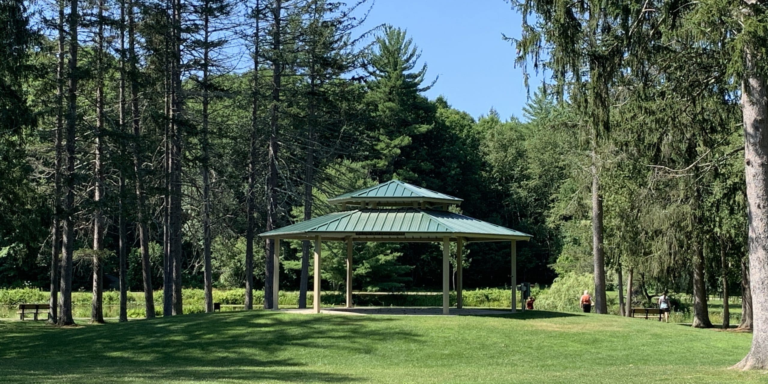 A metal-roofed pavilion stands in a grassy clearing surrounded by tall trees under a clear blue sky, with a few people and benches nearby.