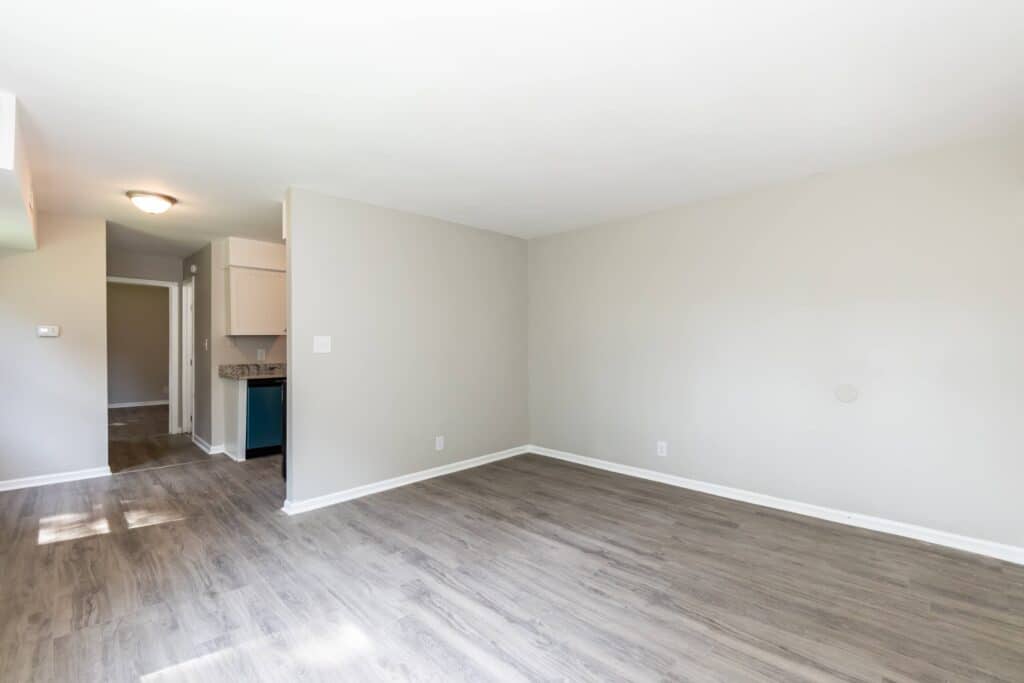 Empty room with light gray walls and wood-look flooring, view of a small kitchen area and hallway in the background under natural and ceiling light.