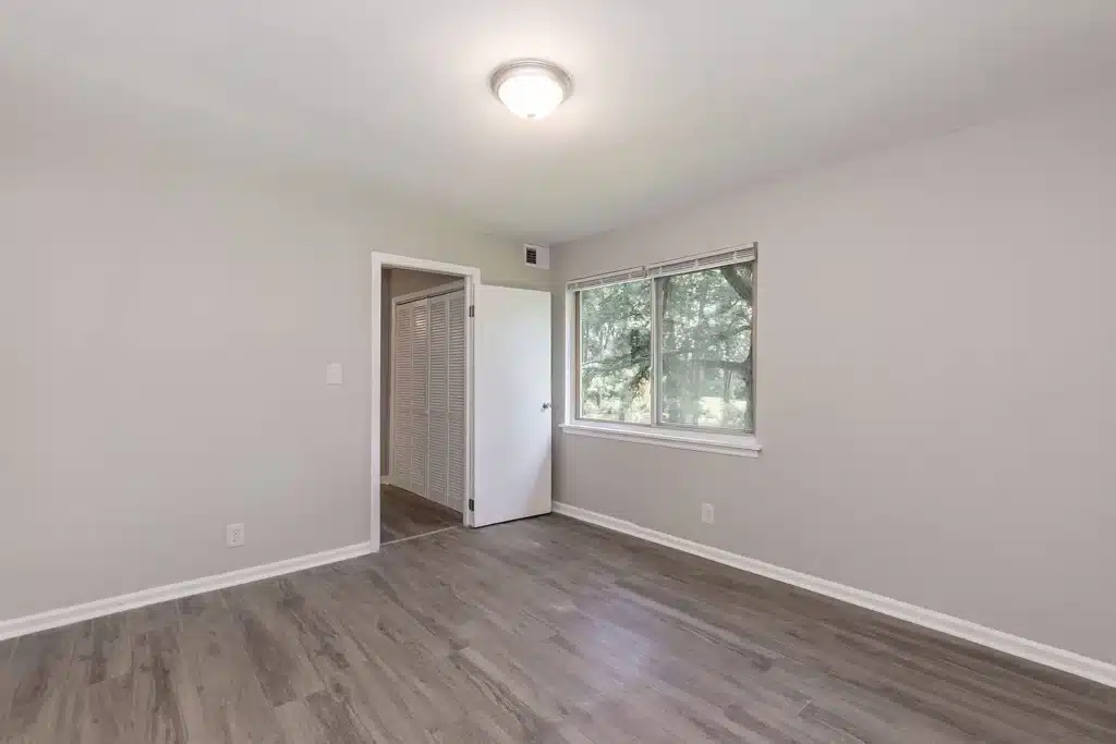 Empty room with light gray walls, wood-style flooring, a ceiling light, an open door, and a window showing trees outside.