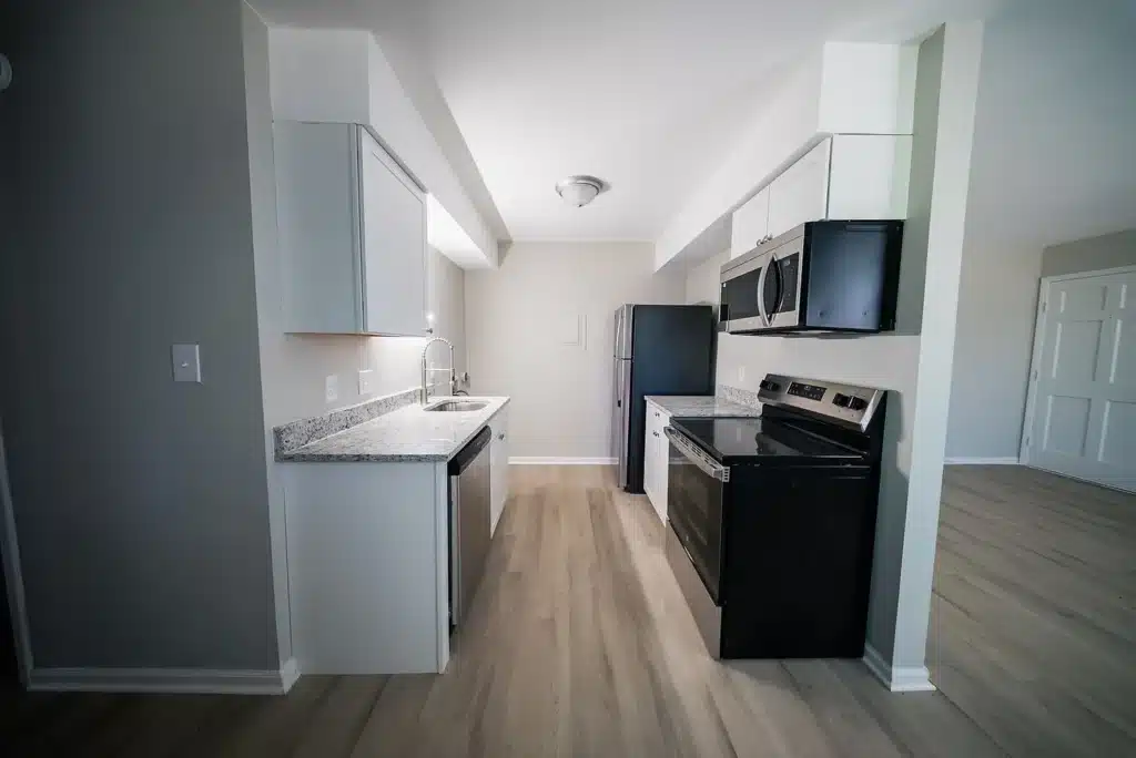 Modern galley kitchen with light wood flooring, white cabinets, granite countertops, stainless steel appliances, and black oven and microwave.