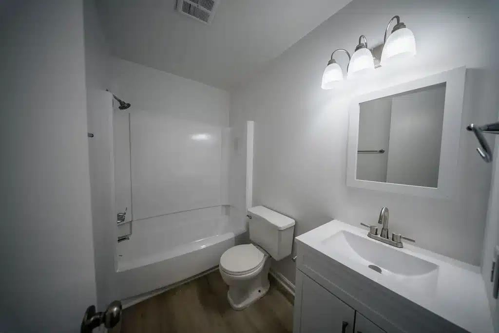 Modern bathroom with a white bathtub and shower, toilet, and white vanity with sink and mirror under three-light fixture.