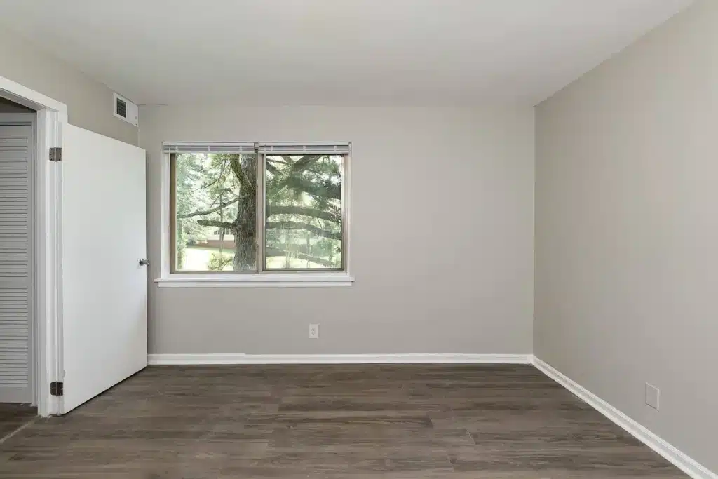 Empty room with light gray walls, a window with blinds showing trees outside, an open white door, and wood-look flooring.