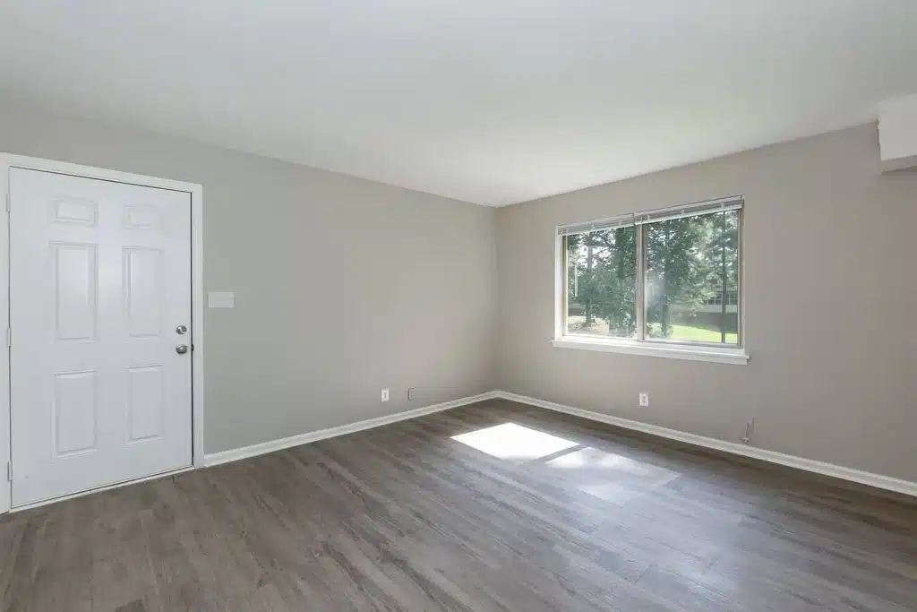 Empty room with light gray walls, wood-style flooring, a white door on the left, and a window on the right letting in natural light.
