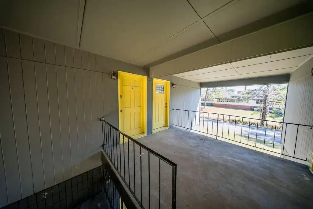 A concrete balcony with black railing features two yellow front doors on a gray-paneled exterior wall, overlooking a street and trees.