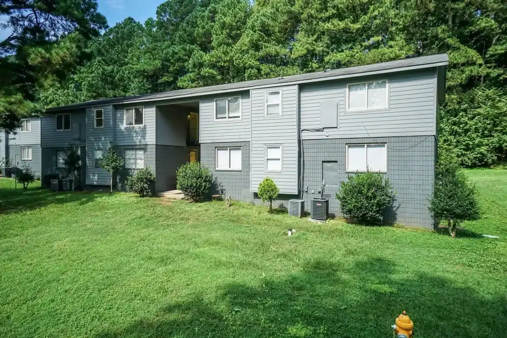 A two-story gray apartment building with several small shrubs in front, set on a grassy lawn with trees in the background.