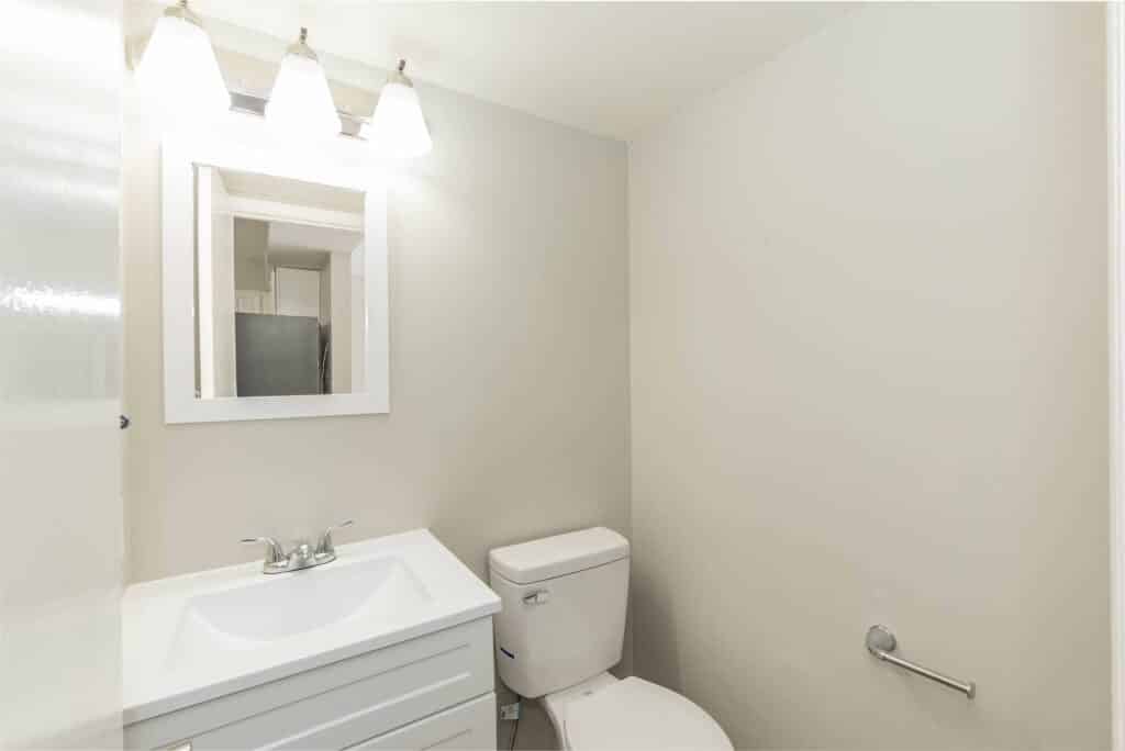 A small bathroom at Gallery - Delmont Townes features a white vanity and sink, a wall-mounted mirror, a toilet, and a three-light fixture above the mirror for bright illumination.