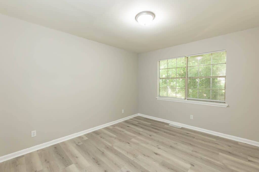 Gallery - Delmont Townes: Empty room with light gray walls, wood-look flooring, a ceiling light, and a window with blinds showing greenery outside.