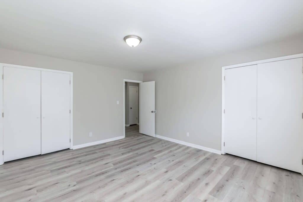 Gallery - Delmont Townes: Empty room with light gray walls, white ceiling, two closed closets, an open door, and light wood flooring under a ceiling light fixture.