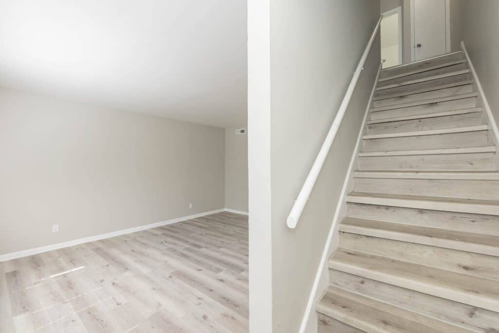 A bright, empty room with light wood flooring sits beside a staircase with matching steps and a white handrail, leading to the hallway above in Gallery - Delmont Townes.