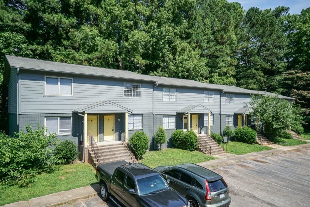 Gallery - Delmont Townes features four gray two-story townhouses with yellow doors, front steps, and small bushes, all set before a wooded area and a parking lot.