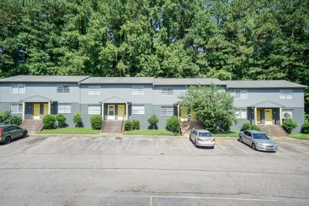 A row of gray, two-story apartment buildings at Gallery - Delmont Townes features steps leading to each entrance, a few parked cars out front, and dense trees in the background.