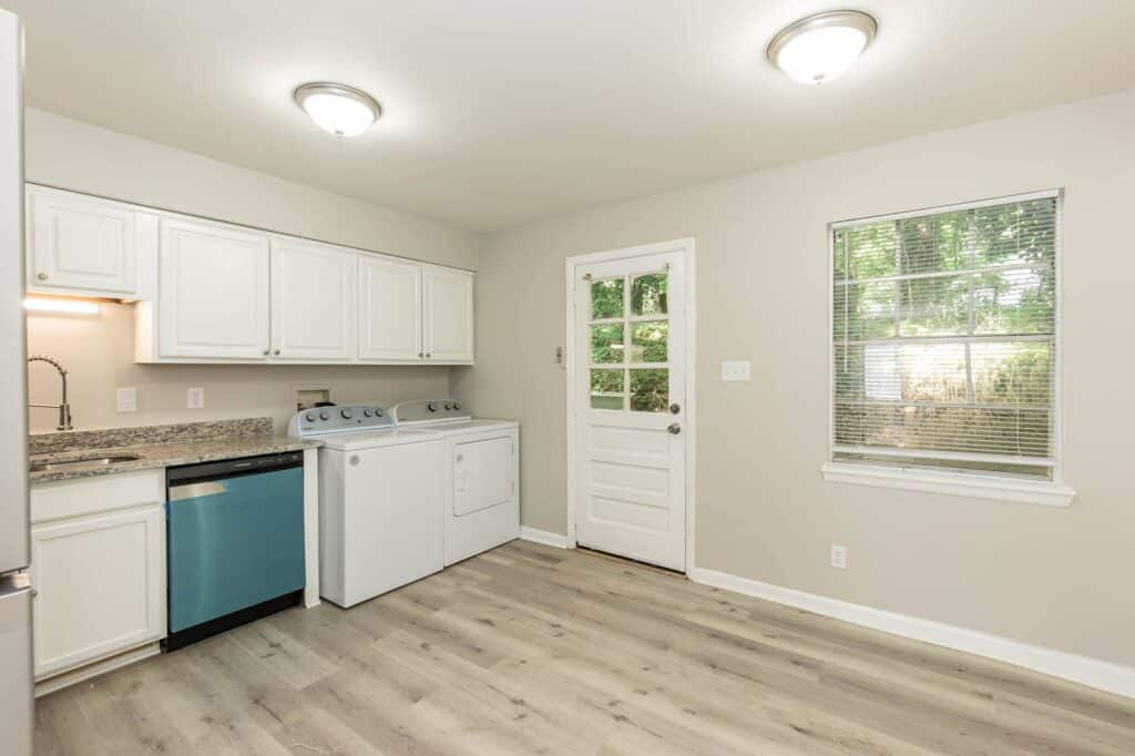 A laundry area in a kitchen at Gallery - Delmont Townes features white cabinets, washer, dryer, stainless steel appliances, wood-style flooring, a door, and a window with blinds.