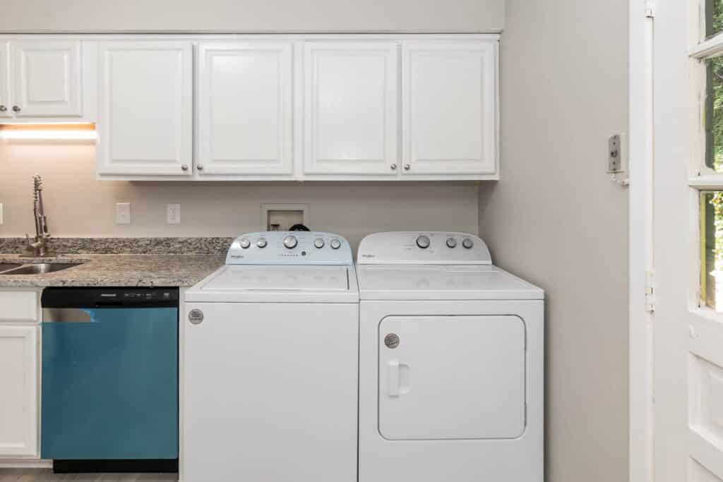 A laundry area at Gallery - Delmont Townes features a white washing machine and dryer side by side, white cabinets above, and a kitchen sink with a blue dishwasher to the left.