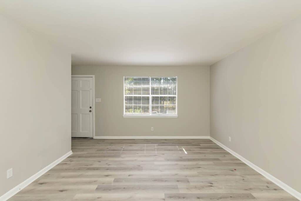 An empty room at Gallery - Delmont Townes features light gray walls, wood-look flooring, a white door on the left, and a window with blinds letting in sunlight.