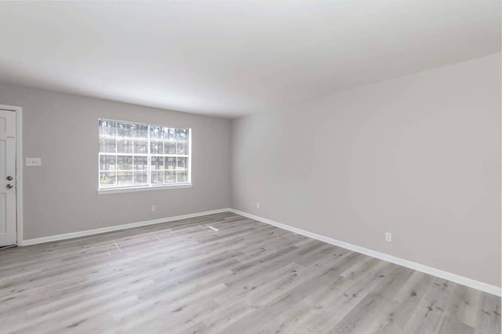 Gallery - Delmont Townes: Empty room with light gray walls, large window with blinds, light wood flooring, and a white door on the left side.