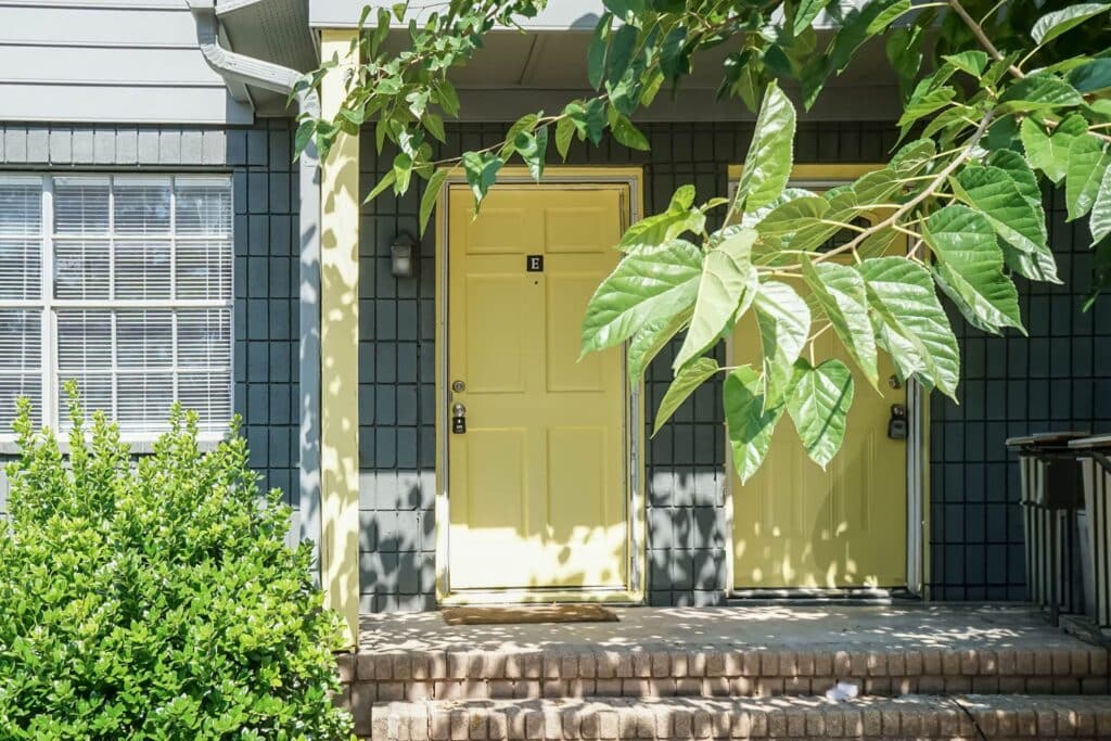 Two yellow doors on the porch of Gallery - Delmont Townes, set against a blue building, are partially shaded by tree leaves. Brick steps lead up, with a window on the left.