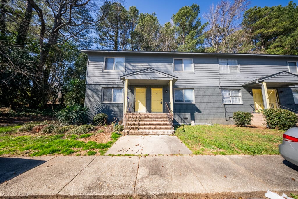 Gallery - Delmont Townes presents a two-story gray townhouse with yellow doors, two front entrances, concrete steps, and a sidewalk, all surrounded by grass, bushes, and trees.
